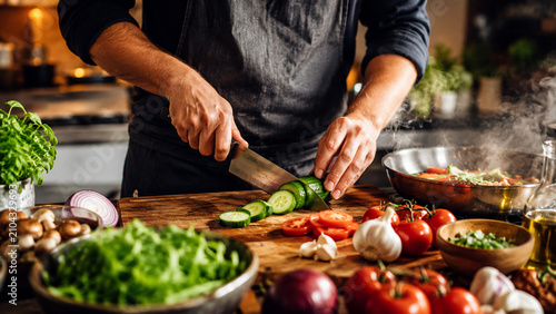 Obraz na płótnie Close‑up of a hand slicing cucumber on a wooden cutting board surrounded by fresh vegetables tomatoes garlic lettuce mushrooms onion and herbs symbolizing home cooking and fresh meal preparation