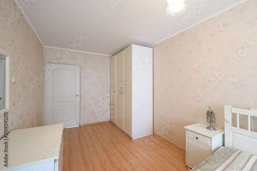 neat bedroom with beige floral wallpaper, a tall cream wardrobe, a white bedside cabinet with a decorative figurine, a wooden floor, and a simple ceiling light