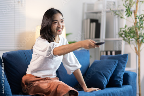 Happy young Asian woman watching TV at home. Young Asian woman smiling, holding a remote control, sitting on a blue couch in her living room, enjoying entertainment.