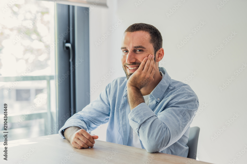Fototapeta premium Smiling young man in a casual shirt sits relaxed at a table by a bright window, conveying confidence, comfort and modern home office lifestyle in natural light.