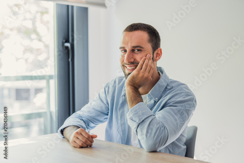 Фотография Smiling young man in a casual shirt sits relaxed at a table by a bright window, conveying confidence, comfort and modern home office lifestyle in natural light