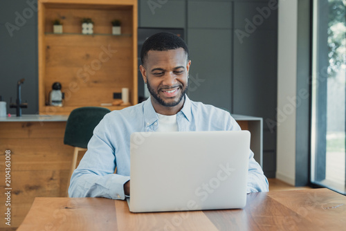 Bild auf Leinwand A smiling young man uses a laptop at a kitchen table, working remotely in a bright, modern home office environment that conveys productivity, comfort, focus and casual professional style