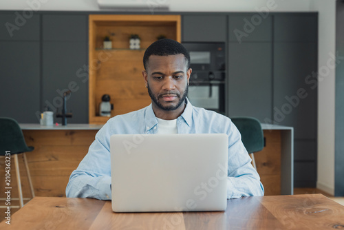 Fotografija A focused young man works on a laptop at a wooden kitchen table, blending home office and remote work in a modern open-plan kitchen while concentrating on tasks or a video call