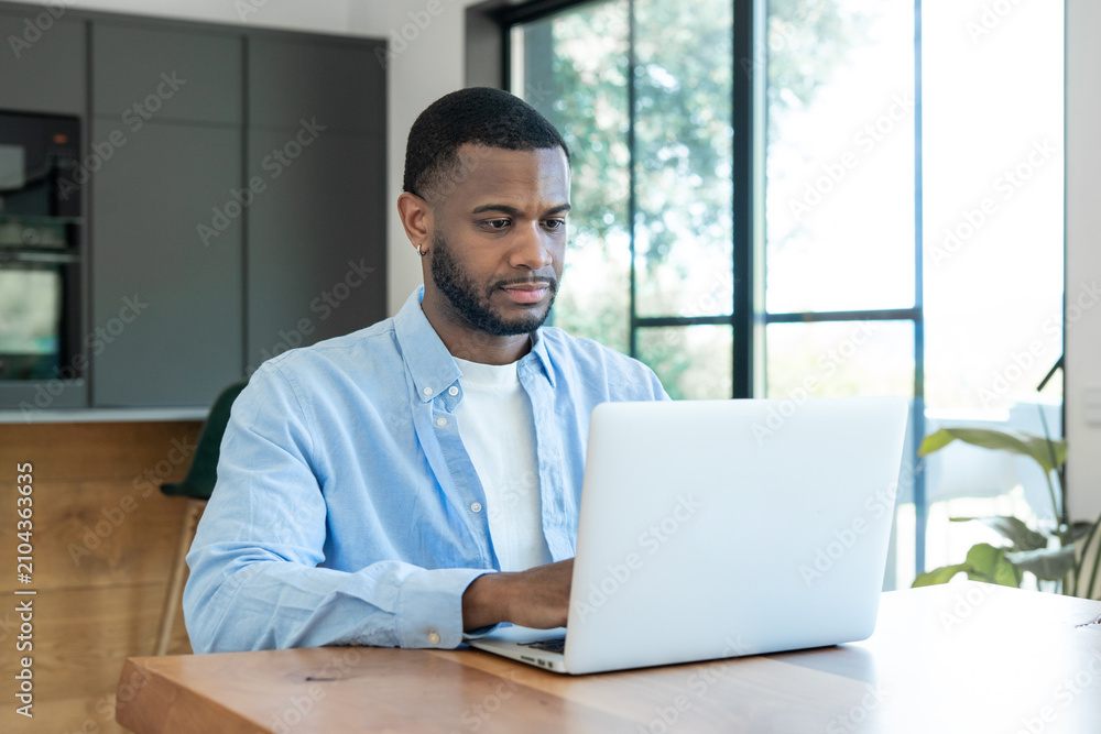 Fototapeta premium Young man seated at a wooden table using a laptop in a bright modern home office, focused on remote work and productivity with natural light from large windows and a casual, professional look.