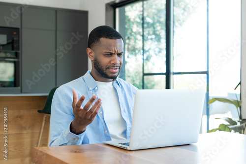 Foto Young man in a bright modern kitchen workspace looks frustrated and confused while using a laptop, reacting to a technical issue during remote work or a video call in a casual home office setting