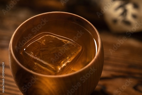 Wooden cup with amber shochu and clear ice cube in soft light.