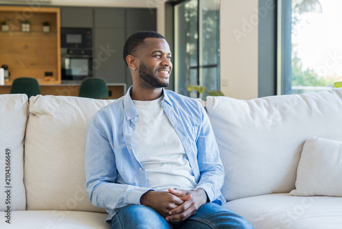 Fotografía Young black man sitting on a comfortable sofa in a modern open-plan living room,