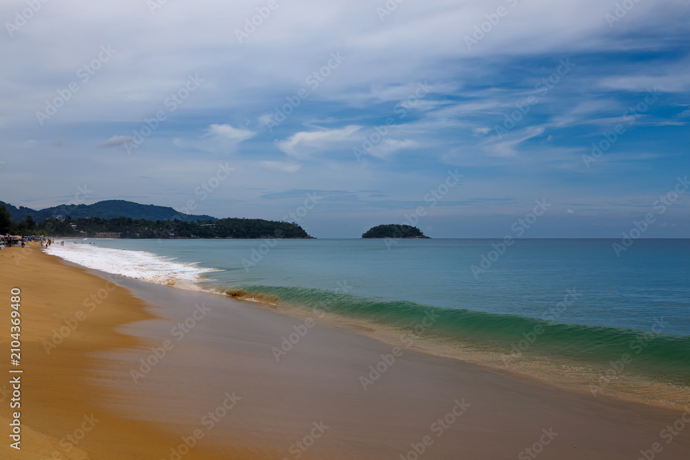 Fototapeta premium Tropical beach view with incoming waves on the sand and a lush green island coastline under cloudy sky