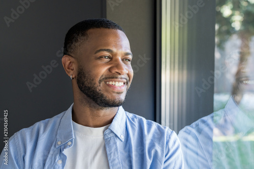 Photos Smiling young man gazes out a window, his reflection visible in the glass; a relaxed indoor portrait conveying happiness, thoughtfulness and warm natural light for lifestyle and business use