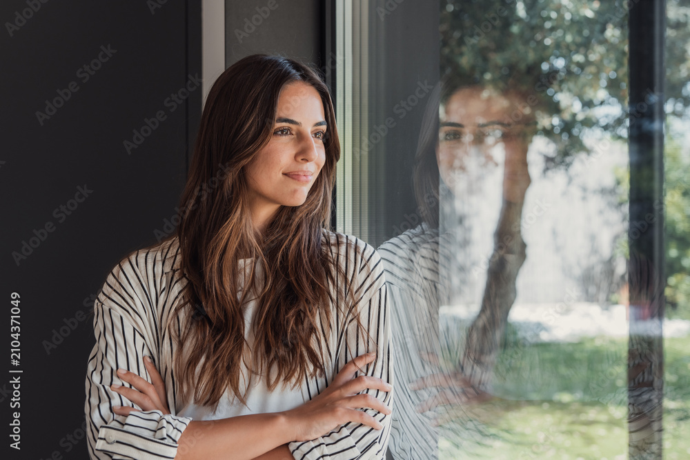 Obraz premium A thoughtful young woman stands with arms crossed, gazing out a window while her reflection appears on the glass, conveying calm, contemplation and modern casual lifestyle in natural light.