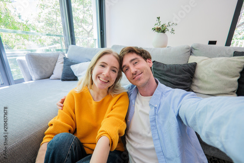 Bild auf Leinwand A happy young couple sits on a comfortable sofa in a bright living room, taking a selfie together
