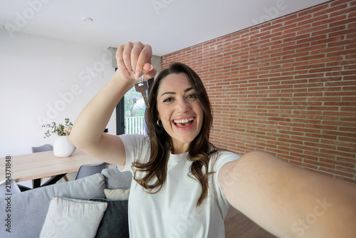 Tableau sur toile A smiling woman celebrates becoming a homeowner as she poses for a selfie holding house keys in a bright, modern apartment living room, conveying excitement, achievement and new beginnings