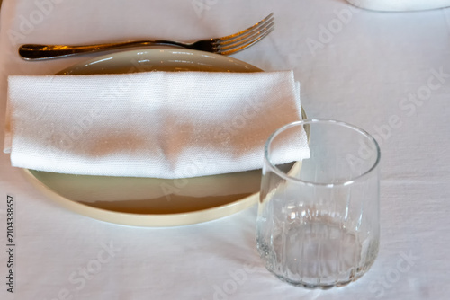 Restaurant table setting with clear drinking glass white cloth napkin and ceramic plate on a white tablecloth
