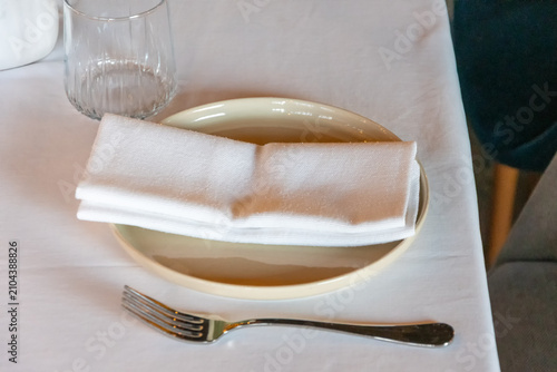 Minimalist restaurant table setting with white cloth napkin fork drinking glass and ceramic plate close up