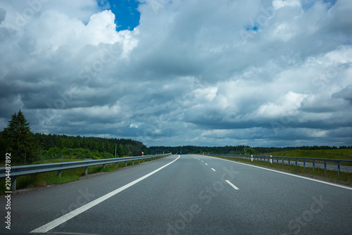 First-person perspective from a car driving on an empty asphalt highway. A modern expressway with a bridge overpass under a dramatic, cloudy stormy sky. No other traffic.
