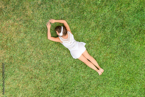 Woman in a white dress and headphones, lying on the green grass sunbathes at summer day. Top view, drone, aerial view