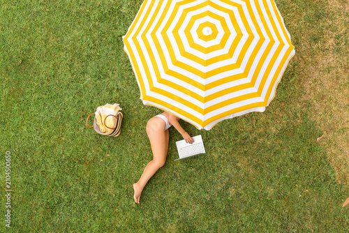 Woman with laptop lying under yellow umbrella on the green grass sunbathes at summer day. Top view, drone, aerial view.