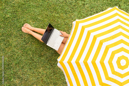 Woman with laptop on the knees sitting on the green grass under yellow umbrella sunbathes at summer day. Top view, drone, aerial view.