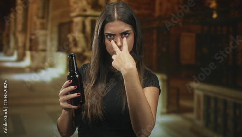 Obraz na plátně Woman holding beer bottle and making v sign over eyes while pointing finger toward camera in building; defiance confrontation