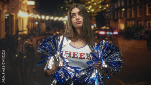 Принти на полотні Young woman cheerleader holding blue metallic pompoms, hands visible, pouting on a street lined with string lights; boredom solitude