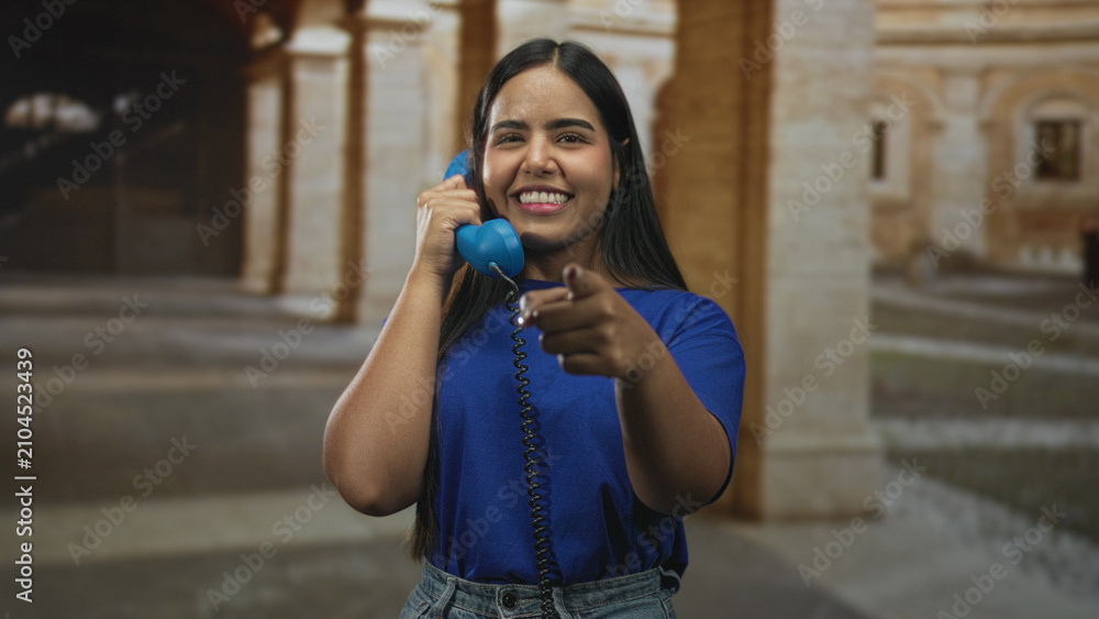 Fototapeta premium Young woman holds blue telephone to her ear and points finger directly toward camera in historic stone building; playful connection joyful.