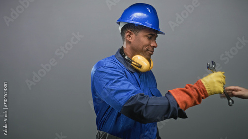 Cuadro en lienzo Man in blue hardhat and work coveralls inspects a wrench with yellow earmuffs and gloved hands in studio; diligence duty