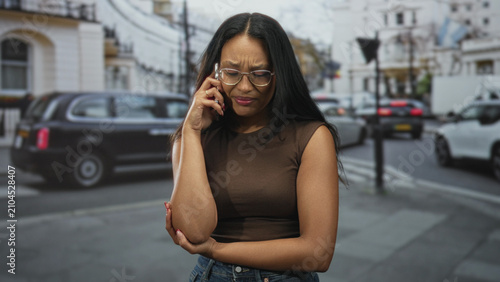 Foto Young hispanic brunette woman wearing glasses and sleeveless top holds smartphone to ear and grips her elbow on a busy city street with cars and buildings; worry