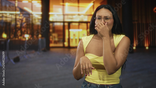 Obraz na plátne Woman pinches nose outside building entrance wearing yellow top and glasses; odor discomfort disgust