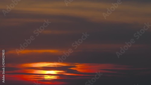 Sun setting behind dark dramatic clouds in a vibrant orange sky during golden hour with a tiny distant airplane flying high above, static wide shot.