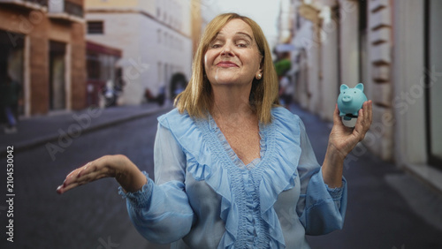 Fotografía Woman holds a blue piggy bank in one hand and shows an open palm with the other on a narrow city street; saving planning hopeful