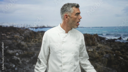 Man in chef jacket looking right on rocky coast with ocean and waves behind, subtle head turn and open mouth gesture; contemplation.