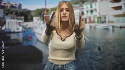 Konstfotografi Woman holding up middle fingers at a harbor pier, redhead in cream sweater and denim jeans with wristwatch visible; defiance attitude