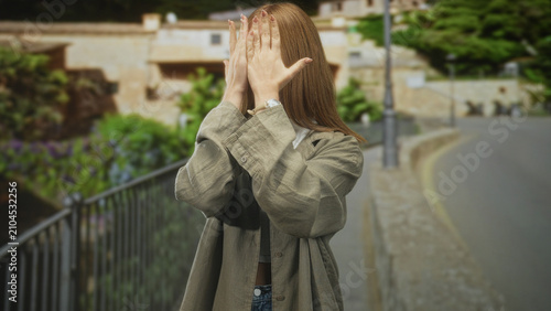 Fotografie Woman with red hair covers face with hands on a narrow stone street by metal railing; shyness privacy