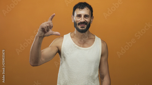 Fotografía Man pinches a small gap with fingers and thumb in a studio orange backdrop, beard and tanktop visible; playful curiosity