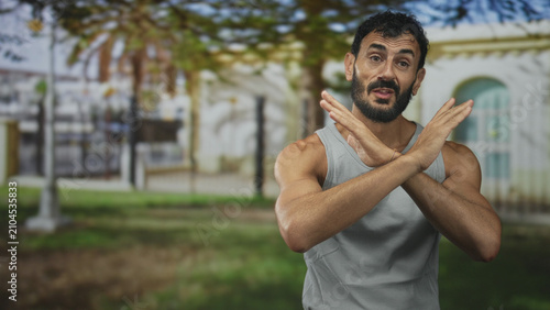 Obraz na płótnie Man with beard crossing forearms in x gesture at building facade, wearing gray tank top and showing muscular arms outdoors; defiance