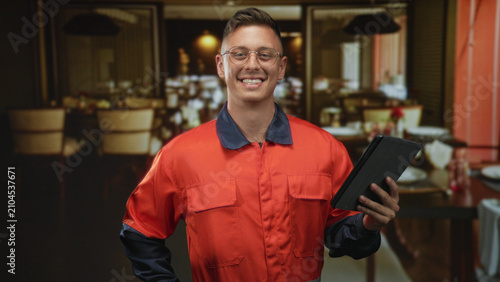Foto Man mechanic in orange coverall holds tablet with right hand, hand on hip, smiling in a restaurant dining room inside building; confidence service