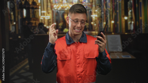 Картината върху платно Man in orange coverall wearing glasses, holding a radio and crossing fingers inside ornate cathedral building; hopeful resilience