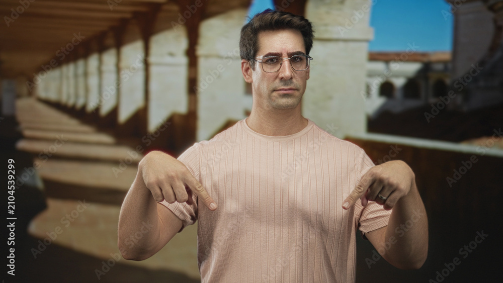 Fototapeta premium Man wearing glasses points both fingers downward at his pink t shirt in a building corridor with stone arches and columns; skepticism instruction.