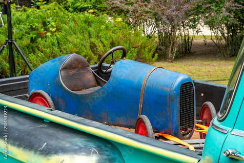 Φωτογραφία Vintage small blue race car soapbox with visible signs of rust and red wheels, placed on the bed of a turquoise pickup truck, outdoor background with greenery
