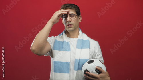 Картината върху платно Man holding soccer ball and rubbing his forehead in studio, wearing blue striped scarf and glasses; disappointment