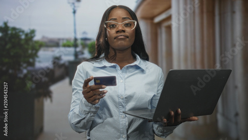 Fotografía Woman holding creditcard in one hand and laptop in the other, glancing skeptically while standing on a street with building columns; financial doubt