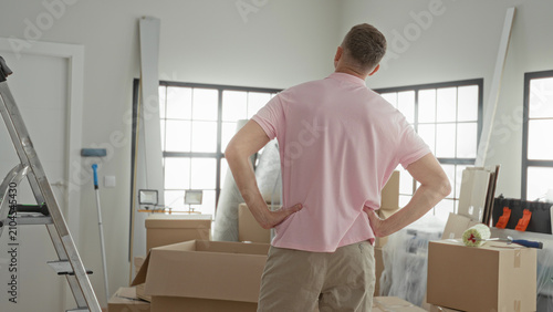 Konstfotografi Man hands on hips inspects stacked moving boxes and a stepladder inside a sunlit building; contemplation planning
