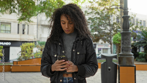 Woman with smartphone scrolling and typing with both hands in street near bench and lamppost  wearing jacket and jeans  focus youth calm.