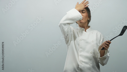 Foto Young woman chef in white coat holds spatula with hand to forehead and open palm gesture in studio; frustration
