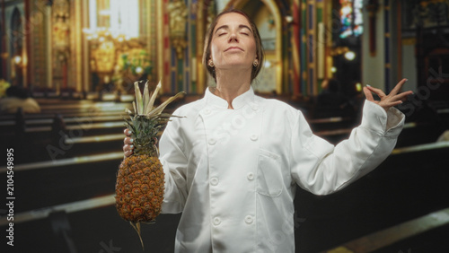 Obraz na plátne Woman chef holding pineapple with meditative hand gesture and closed eyes in a building; culinary ritual serenity