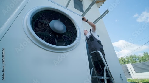HVAC technician on a ladder installing a large outdoor heat pump unit on a modern house wall.