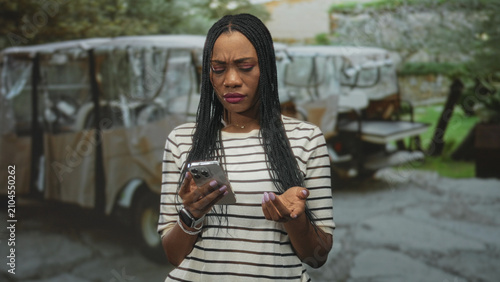 Fotografie Black woman holding smartphone and frowning while checking phone on street, wearing striped top and smartwatch; frustration solitude