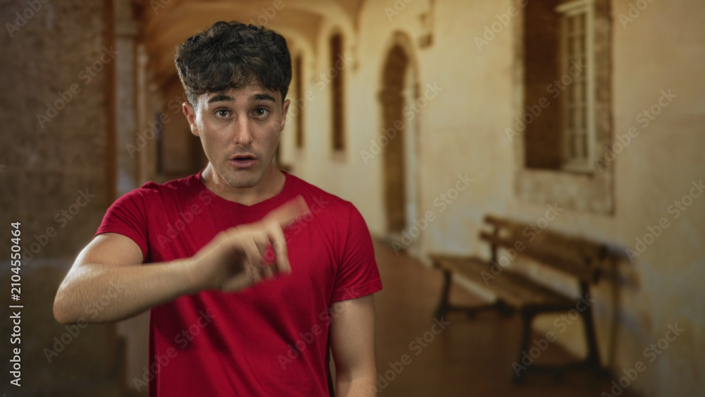 Obraz premium Man in red shirt gesturing with hand in building corridor, speaking and frowning; uncertainty reflection.