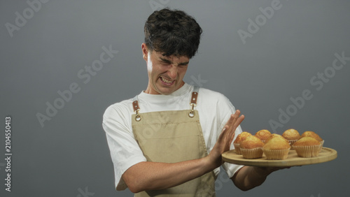 Fotografía Young hispanic man in beige apron pushes away a wooden tray of muffins while grimacing in a gray studio; disgust refusal avoidance
