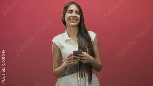 Fotografía Young woman holds smartphone with both hands in studio, smiling and glancing upward, long hair and white blouse visible; playful curiosity